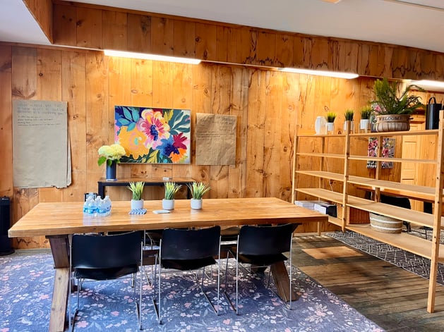 "A wood-paneled coworking meeting room with a long communal table, black chairs, a floral rug, and open shelving styled with plants and decor."