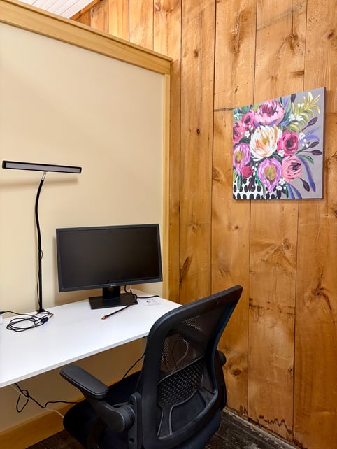A cozy private office with decorative cedar planks, floral artwork, and a dedicated professional workstation with chair, adjustable desk, monitor, and task lighting. 
