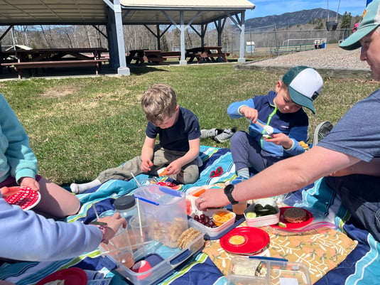 Family picnicking on a blanket at the Carrabassett Valley Town Playground in Maine, with covered pavilions and picnic tables in the background