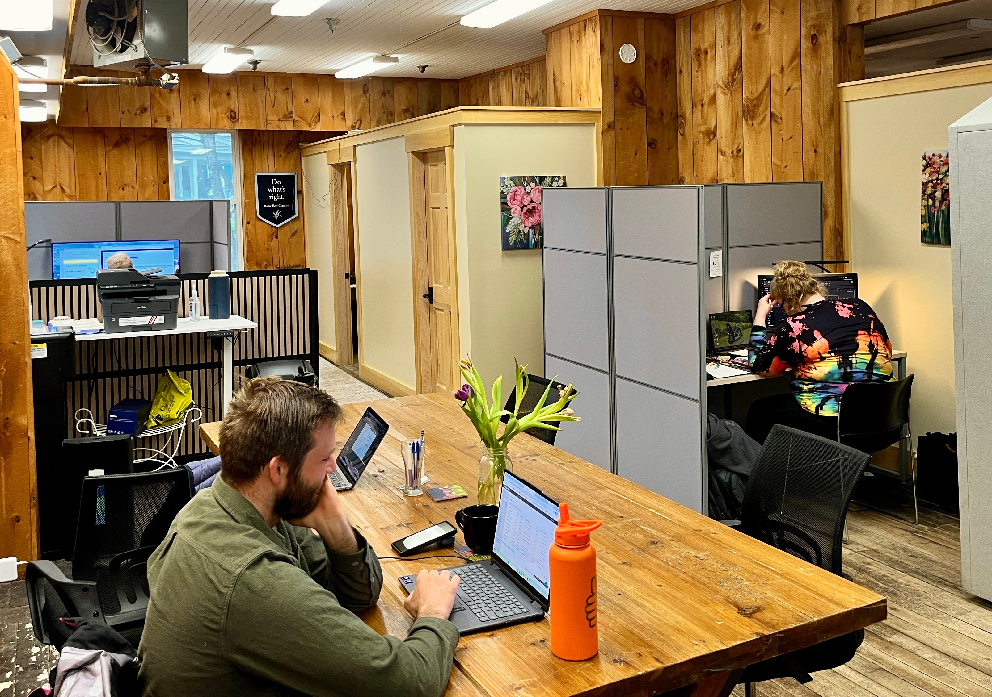 "Two coworking members working at a shared wooden table and private cubicles in a wood-paneled office space, with a third member on a call in the background."