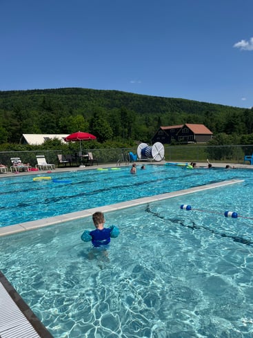 Image (Carrabassett Valley Public Pool): Child swimming in the outdoor Carrabassett Valley Public Pool in Maine on a sunny summer day, with wooded scenery in the background