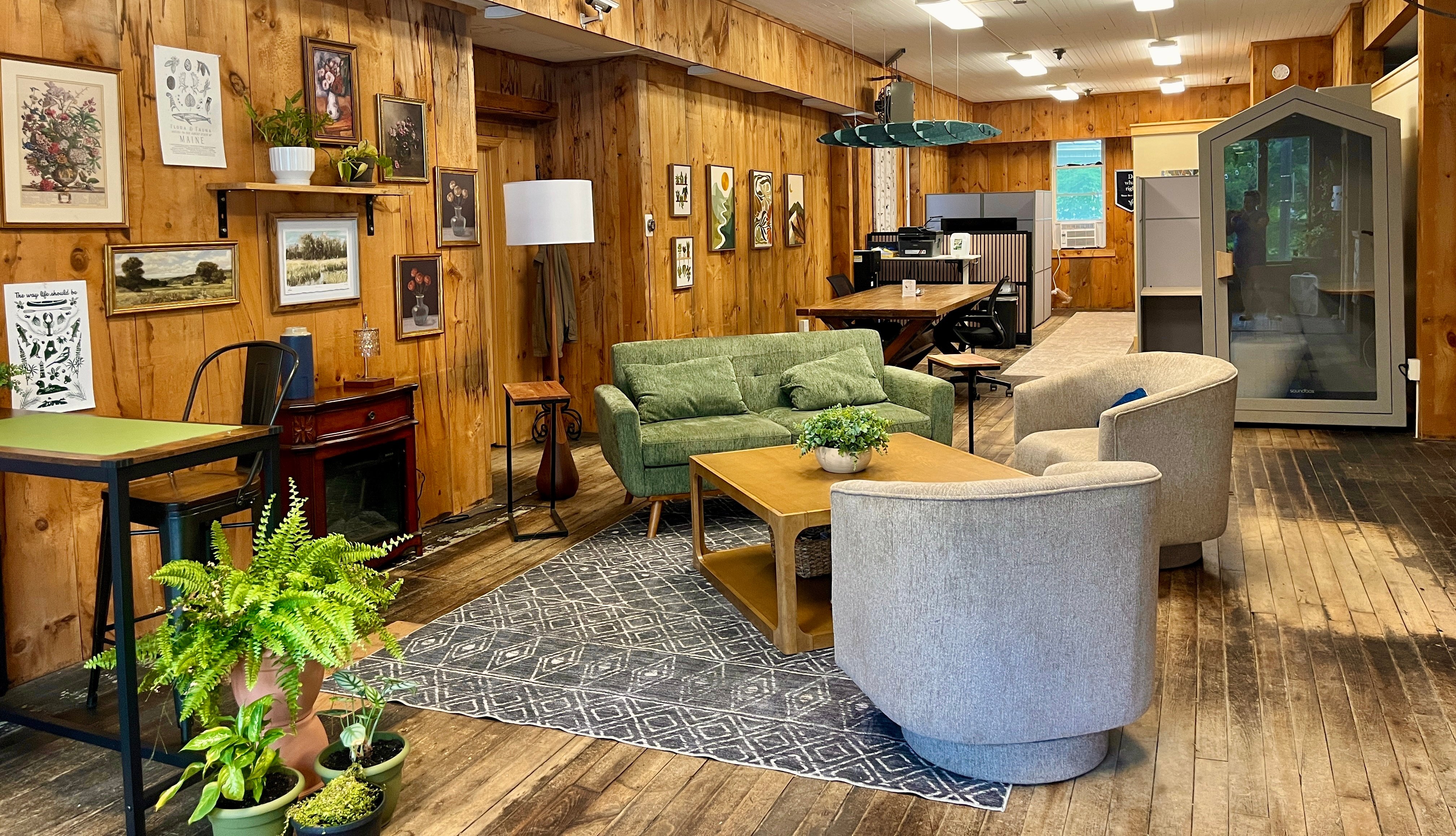 A cozy wood-paneled lounge area with a green sofa, gray armchairs, a wooden coffee table, potted plants, and a gallery wall of framed art and Maine-themed prints."