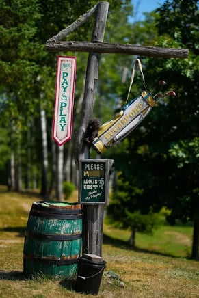 Honor-system "Please Pay to Play" sign and barrel at Moose Meadows Golf Course, a $10 par-3 wilderness golf course in Stratton, Maine